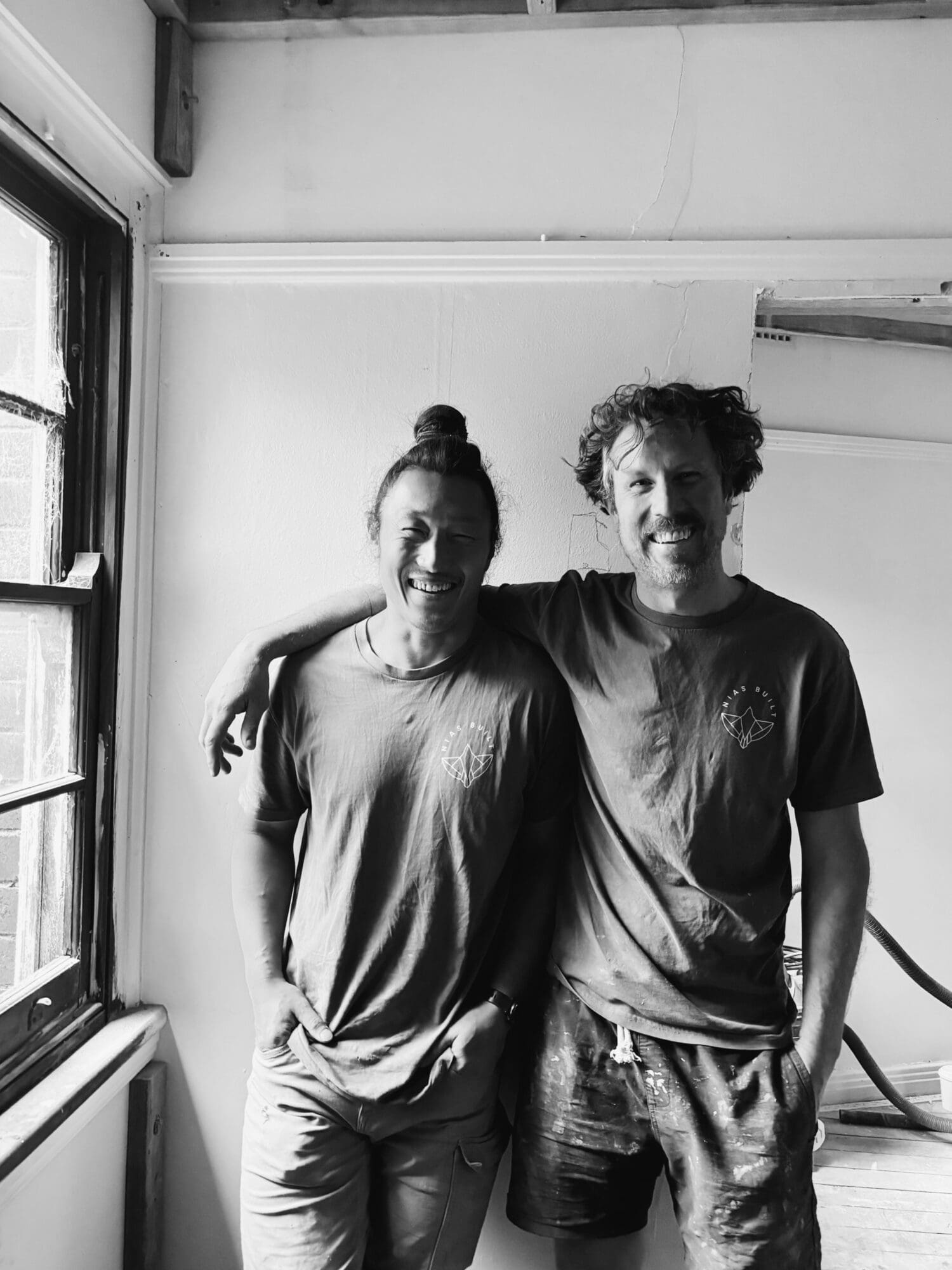 Two smiling men, one with an arm around the other, in a room under renovation